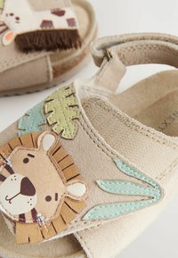 Helle beigen Sandalen mit einer Löwenkopf-Applikation, grünen Blättern, einem weichen Textilriemen und einem strukturierten Fußbett. Mit Fransen-Details am Riemen.