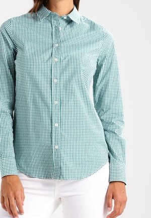Frau trägt ein eng anliegendes grün-weiß kariertes Hemd mit Knöpfen, langen Ärmeln und weiße Hose vor einfachem Hintergrund.