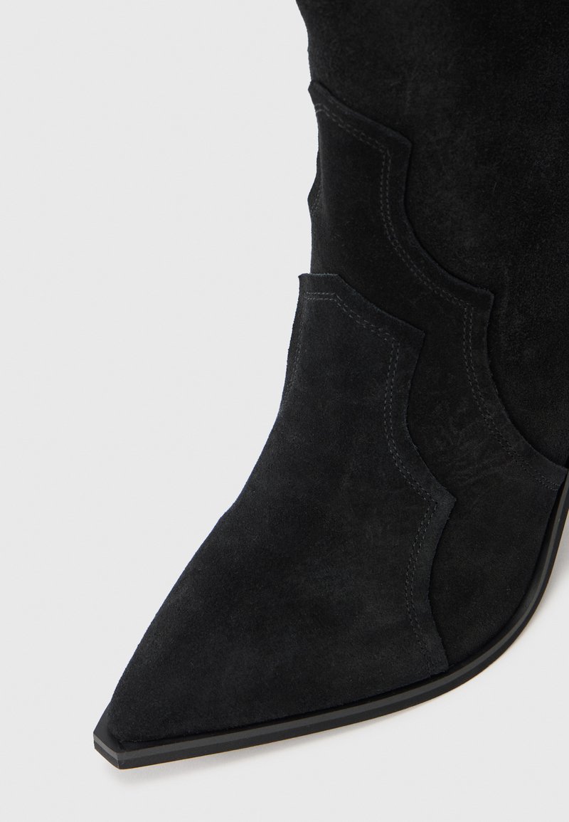 Stivale alla caviglia in pelle scamosciata nera con punta affusolata, caratterizzato da cuciture decorative e ritagli arcuati lungo il lato superiore. Design conciso e strutturato.