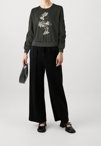 Sweat-shirt vert foncé avec un motif de personnage de dessin animé, associé à un pantalon large noir et un petit sac noir. Des chaussures plates noires complètent la tenue.