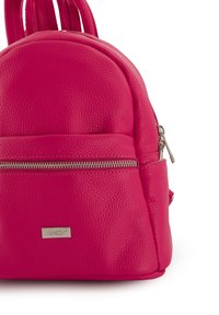 Sac à dos en cuir rose avec une forme arrondie, surface texturée, poche zippée à l'avant et plaque en métal de marque. Deux courtes poignées de transport.