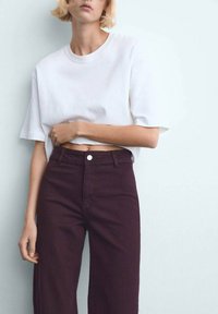 Mlada oseba s kratkimi blond lasmi, oblečena v preprosto belo cropped majico in temno vijolične hlače z visokim pasom, stoji ob svetlem zidu.