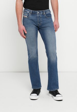 Homme portant un jean droit bleu moyen, des baskets noires et blanches, debout devant un fond clair uni.