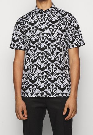 Man wearing short-sleeve black shirt with bold white symmetrical abstract pattern, paired with black pants, standing against plain background.