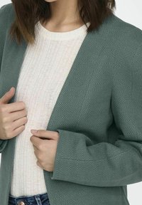 Veste verte à motif chevrons avec une finition texturée ; portée sur un pull léger à côtes. La tenue présente une coupe décontractée et des manches ajustées.