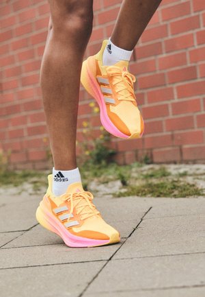 Osoba nosící oranžové a růžové běžecké boty Adidas s bílými ponožkami Adidas, zachycena uprostřed kroku na dlážděném povrchu poblíž cihlové zdi.