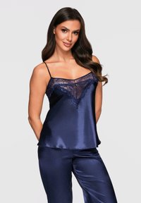 Kvinna iklädd en marinblå satin camisole med spetstillbehör och matchande satinbyxor, stående mot en enkel ljus bakgrund.