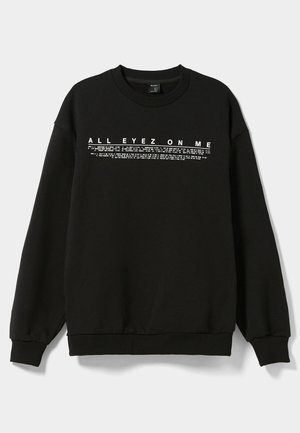 Person trägt schwarzen Sweatshirt mit weißem Schriftzug "ALL EYEZ ON ME" und kleinen bedruckten Linien, kombiniert mit glänzenden silbernen Hosen.