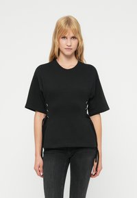 Μαύρο κοντομάνικο t-shirt με άνετη εφαρμογή, με κορδόνια στα πλαϊνά με μεταλλικά κουμπώματα, κατασκευασμένο από λείο ύφασμα.