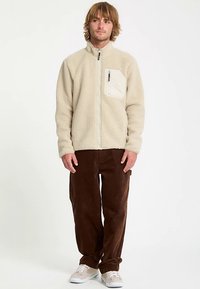 Béžová fleecová bunda s vysokým golierom, predným zipsom a ľavým náprsným vreckom; nosená s hnedými kordurovými nohavicami a teniskami.