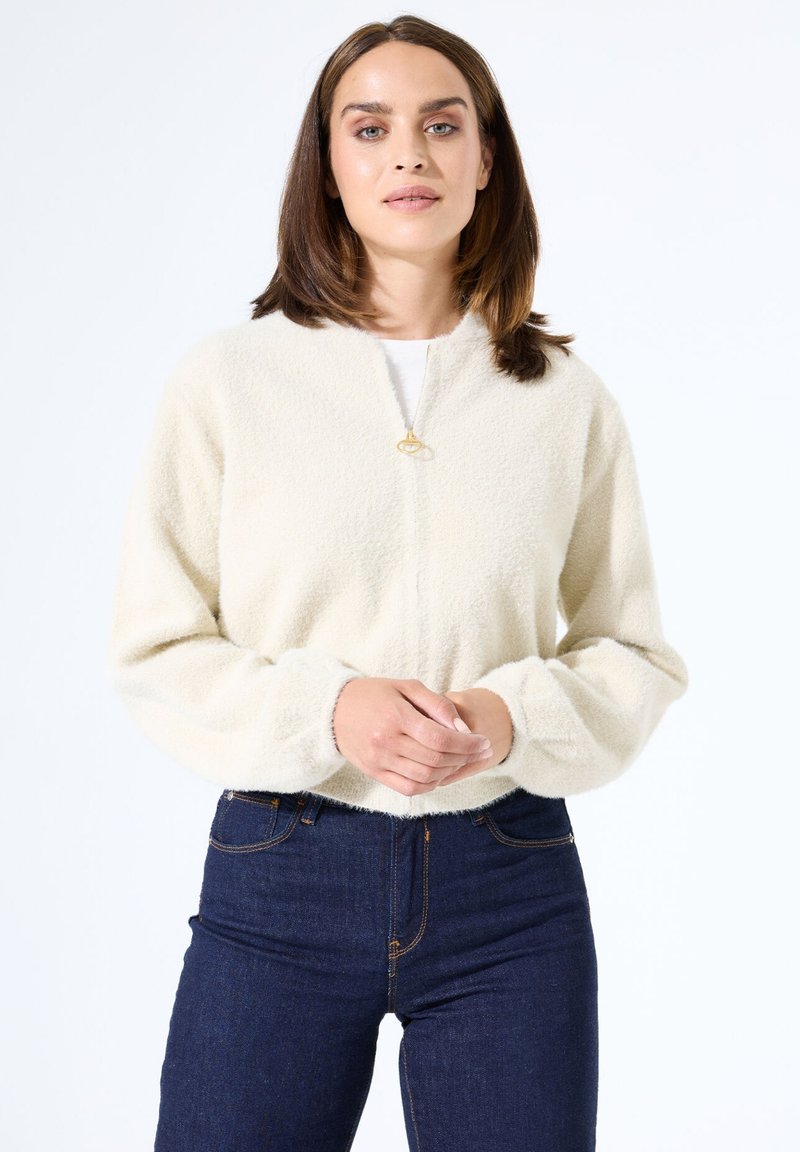 Cremiger, flauschiger Pullover mit rundem Halsausschnitt und Reißverschluss. Kombiniert mit dunkelblauen, hoch taillierten Jeans mit sichtbaren Nähten.