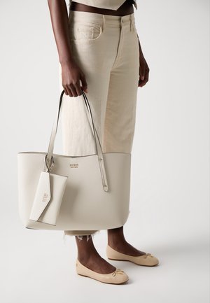 Person wearing cream pants and beige flats holding a large textured white Guess tote bag with matching small pouch.