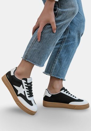 Personne portant un jean bleu raccourci et des baskets noir et blanc avec un motif étoile, levant légèrement un pied du sol.