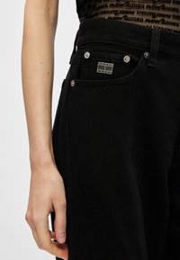 Jeans de ganga preta de cintura alta, com fechos em metal prateado, um bolso frontal com um etiqueta "Tommy Jeans" e um acabamento de tecido texturizado.