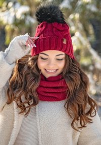 Gorro de lana rojo trenzado con un pompón negro, que presenta un diseño acanalado. Acompañado de una bufanda de lana roja a juego. Tela suave y texturizada.