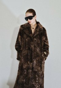 Cappotto in finta pelliccia marrone con motivi a onda testurizzati, grande colletto a punta e bottoni frontali. Il modello indossa occhiali da sole oversize neri e una collana.