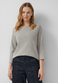 Vrouw met golvend blond haar, gekleed in een lichtgrijze gebreide trui en donkere high-waisted broek, staande tegen een eenvoudige lichte achtergrond.