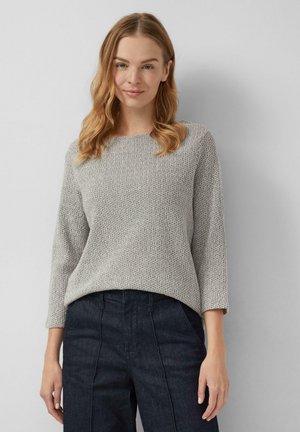 Frau mit welligem blonden Haar, die einen hellgrauen strukturierten Pullover und dunkle hochgeschnittene Hosen trägt, steht vor einem schlichten, hellen Hintergrund.