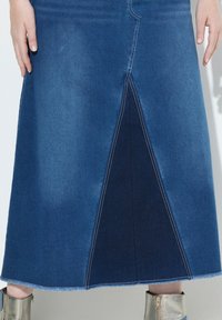 Falda de mezclilla con una abertura frontal, que presenta tonos variados de azul, con dobladillos terminados y costuras en contraste. Se lleva con botas metálicas hasta el tobillo.