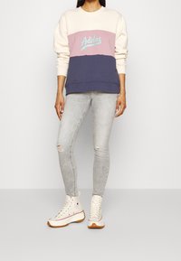 Persona che indossa una felpa Adidas tricolore, jeans skinny strappati grigio chiaro e sneaker alte bianche con suola marrone.