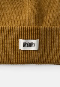Bonnet tricoté brun avec une texture à côtes verticales, arborant une étiquette blanche portant le texte "DRYKORN" en lettres noires en gras.