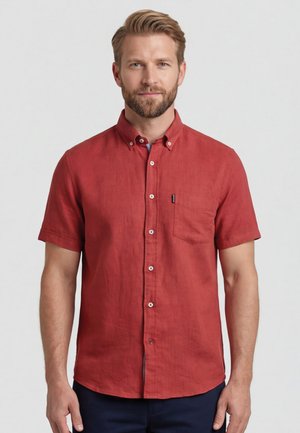 Man met lichte baard en blond haar, die een rood knoopovershirt met korte mouwen en een borstzakje draagt, tegen een effen achtergrond.