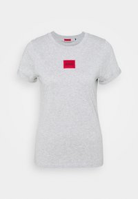 Grå bomulds-t-shirt med optrukne ærmer, med en rød firkantet lap med "HUGO" label i midten. Glat tekstur, afslappet pasform.