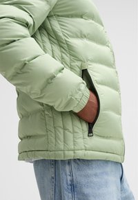 Giacca a vento verde chiaro con cuciture orizzontali, tasca laterale con zip e polsini a coste. Il tessuto appare liscio e imbottito.