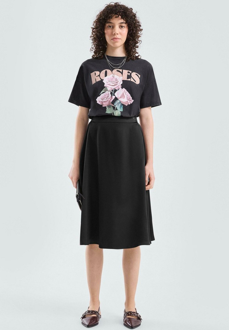 Czarny T-shirt z nadrukiem róży i napisem "ROSES", zestawiony z czarną spódnicą w kształcie litery A. Modelka ma na sobie brązowe mokasyny oraz warstwowe srebrne naszyjniki.