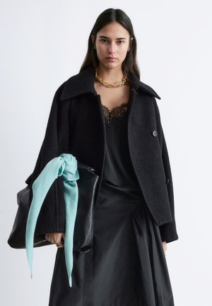 Frau mit langem dunklem Haar, die einen schwarzen Mantel über einem schwarz mit Spitze besetzten Kleid trägt, eine goldene Kettenkette um den Hals und eine schwarze Ledertasche mit einem an den Griff gebundenen blauen Schal in der Hand hält.