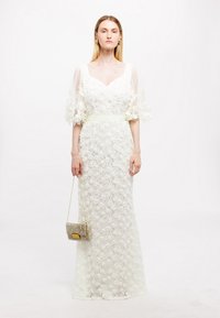 Witte bloemenkant jurk met doorschijnende, uitlopende mouwen, een aangesloten taille en een rechte silhouet. Accessoires zijn een kleine, versierde handtas.