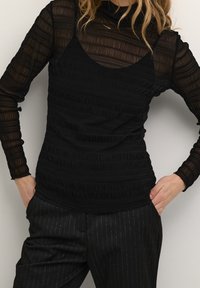 Svart långärmad topp i ett strukturerat genomskinligt meshmaterial med ett åtsittande underlag, tillsammans med svarta randiga byxor.