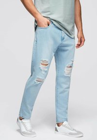 Jeans de mezclilla azul claro con desgarros desgastados en las rodillas, pierna ajustada, diseño de cinco bolsillos, combinados con zapatillas deportivas blancas y grises.