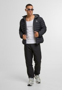 Chaqueta de plumas negra con capucha, diseño acolchado horizontal, combinada con pantalones jogger grises y una camiseta sin mangas blanca. Mínima marca en la chaqueta.
