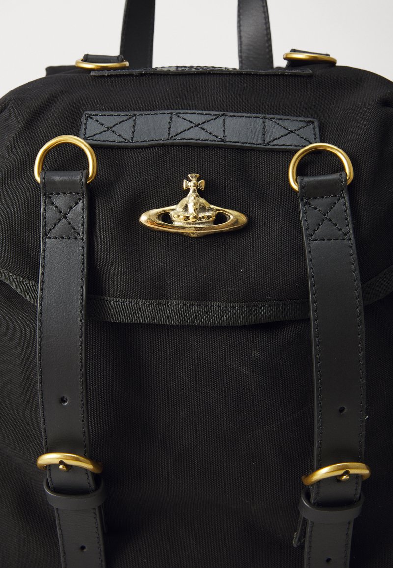 Zaino in tela nera con cinturini in pelle, hardware in metallo dorato e un emblema decorativo a forma di corona. Presenta dettagli in pelle texturizzata.
