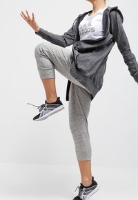 Persona en ropa atlética gris y zapatillas negras levantando una pierna, brazos extendidos, contra un fondo blanco liso.