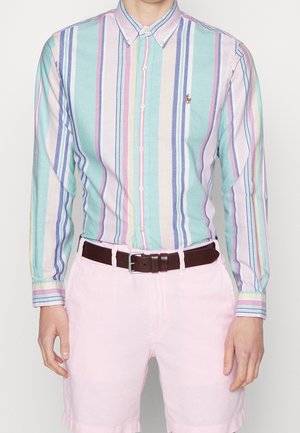 Mann trägt ein pastellgestreiftes Langarmhemd, das in hellrosa Shorts mit einem dunkelbraunen Gürtel gesteckt ist.
