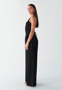 Black jumpsuit with thin straps, fitted bodice, and wide-leg pants. Smooth fabric, features a drawstring detail at the back.