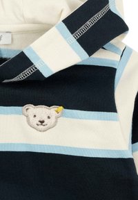 Gestreifte Kapuzenjacke in Marineblau, Creme und Hellblau. Mit einem gestickten Bärenkopf auf der Brust und genähten Details an den Kanten.