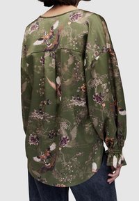 Blouse à manches longues, verte, avec un motif floral et d'oiseaux. Présente une coupe décontractée, un col rond et des poignets froncés. Texture de tissu lisse.