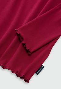 Chemise bordeaux à manches longues avec une texture côtelée. Présente des poignets volantés et une étiquette de marque visible étiquetée "TOM TAILOR."