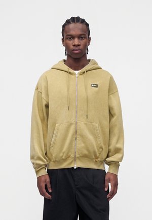 Giovane uomo con capelli intrecciati indossa una felpa beige con zip e logo Nike e pantaloni neri, in piedi davanti a uno sfondo bianco uniforme.