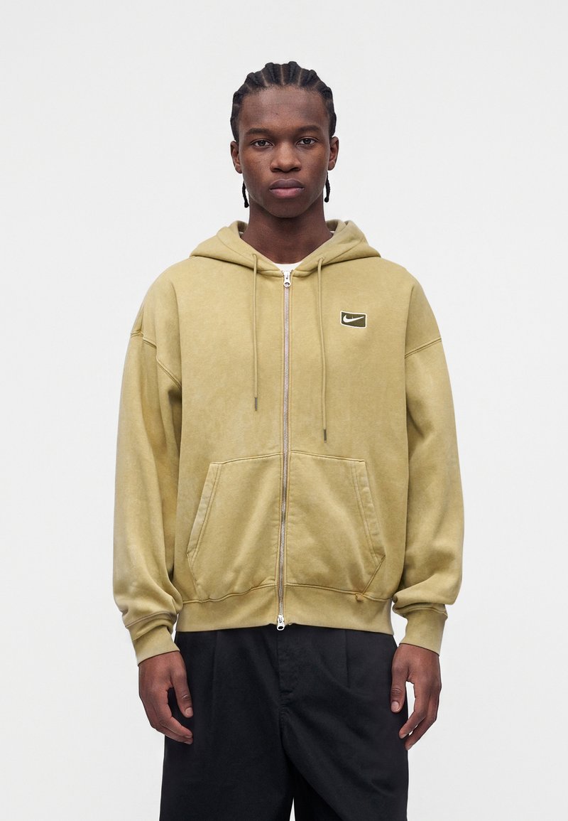 Junger Mann mit geflochtenem Haar trägt einen beigen Hoodie mit Reißverschluss und Nike-Logo sowie schwarze Hose und steht vor einem schlichten weißen Hintergrund.