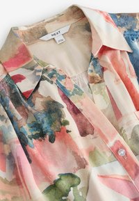 Blouse à col avec une fermeture à boutons sur le devant, arborant un imprimé floral aquarelle dans des tons de rose, bleu, vert et beige.