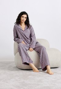 Conjunto de pijama a rayas en tonos morado y rosa, con mangas largas y pantalones, colocado sobre una silla texturizada de color claro en un entorno neutral.