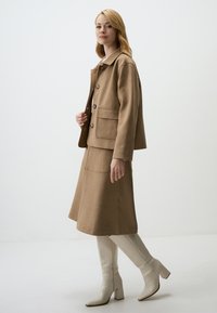 Giacca in misto cotone marrone con chiusura a bottoni, tasche laterali, abbinata a una gonna a ginocchio coordinata e stivali bianchi con tacco a blocco al ginocchio.
