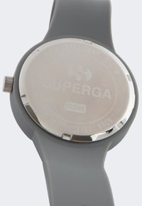 Reloj de silicona gris con una parte trasera redonda de acero inoxidable, que presenta el logotipo de la marca grabado y el detalle de resistencia al agua. Textura suave y mate.
