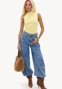 Femme portant un top jaune sans manches froncé, un jean bleu à jambes larges avec des pierres colorées, des bottes en daim marron et tenant un sac à main marron duveteux.