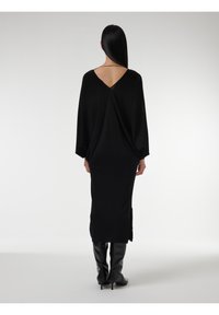 Zwarte gebreide jurk met V-back, lange mouwen, relaxte pasvorm, zijsplitten. Gepaard met knie-high zwarte stiletto laarzen. Glad textuur.