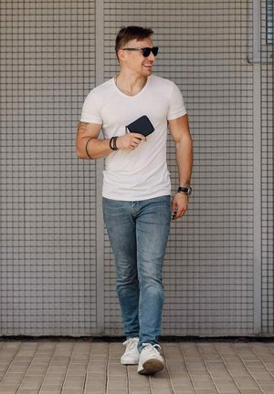 Bílé tričko, modré džíny a bílé tenisky. Drží černou peněženku. Brýle na slunce, jde proti pozadí se vzorem mřížky.